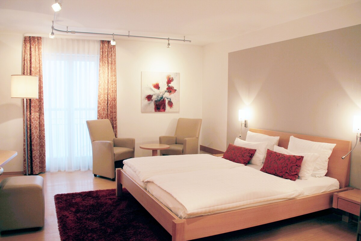 A hotel room with a bed, two chairs, a small table, a lamp, and a painting on the wall.