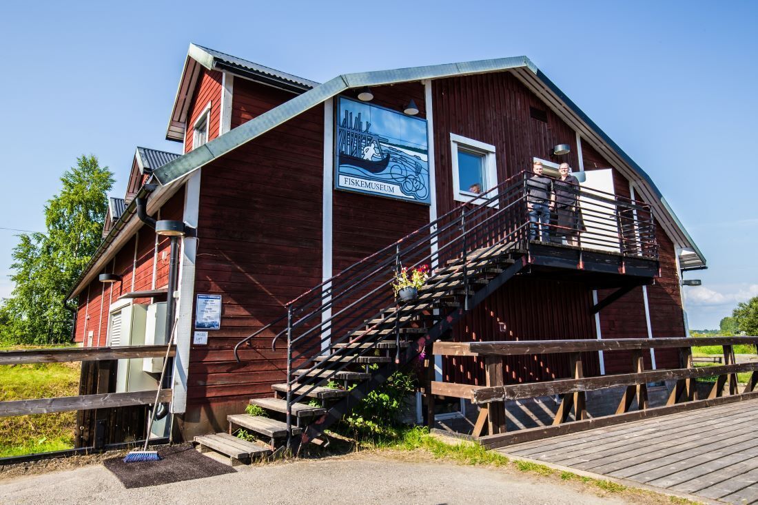 Un bâtiment en bois avec un escalier menant à un balcon, une enseigne indiquant « FISKMUSEUM », et une petite fenêtre.