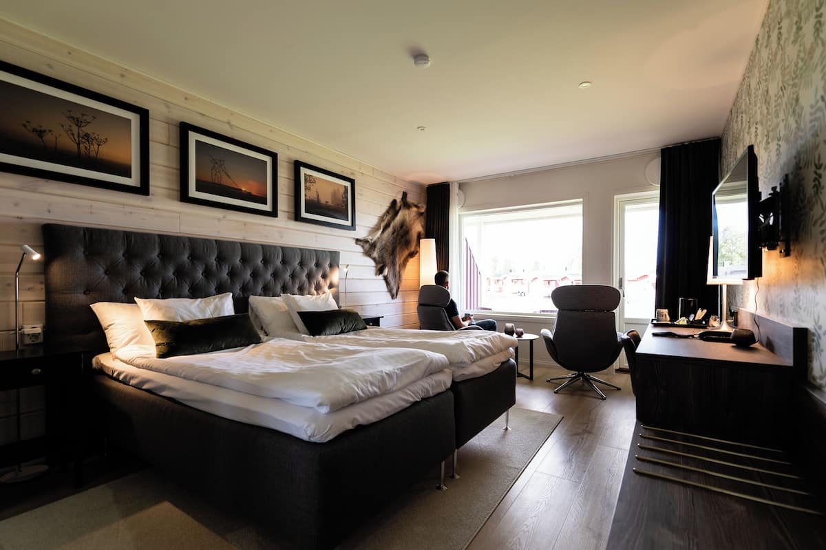 Une chambre d’hôtel moderne avec un grand lit, un bureau et une chaise. Les murs sont ornés de tableaux encadrés et il y a une fenêtre avec des rideaux.