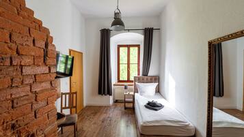 A room with a brick wall, a television, a bed, a chair, a window with curtains, and a mirror.