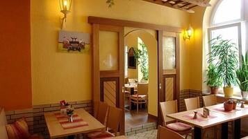 A restaurant interior with wooden tables, chairs, and a brick wall. There are potted plants and a painting on the wall.