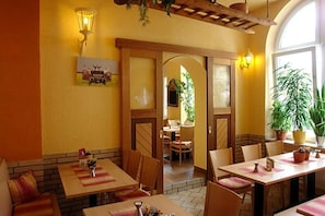 A restaurant interior with wooden tables, chairs, and a brick wall. There are potted plants and a painting on the wall.