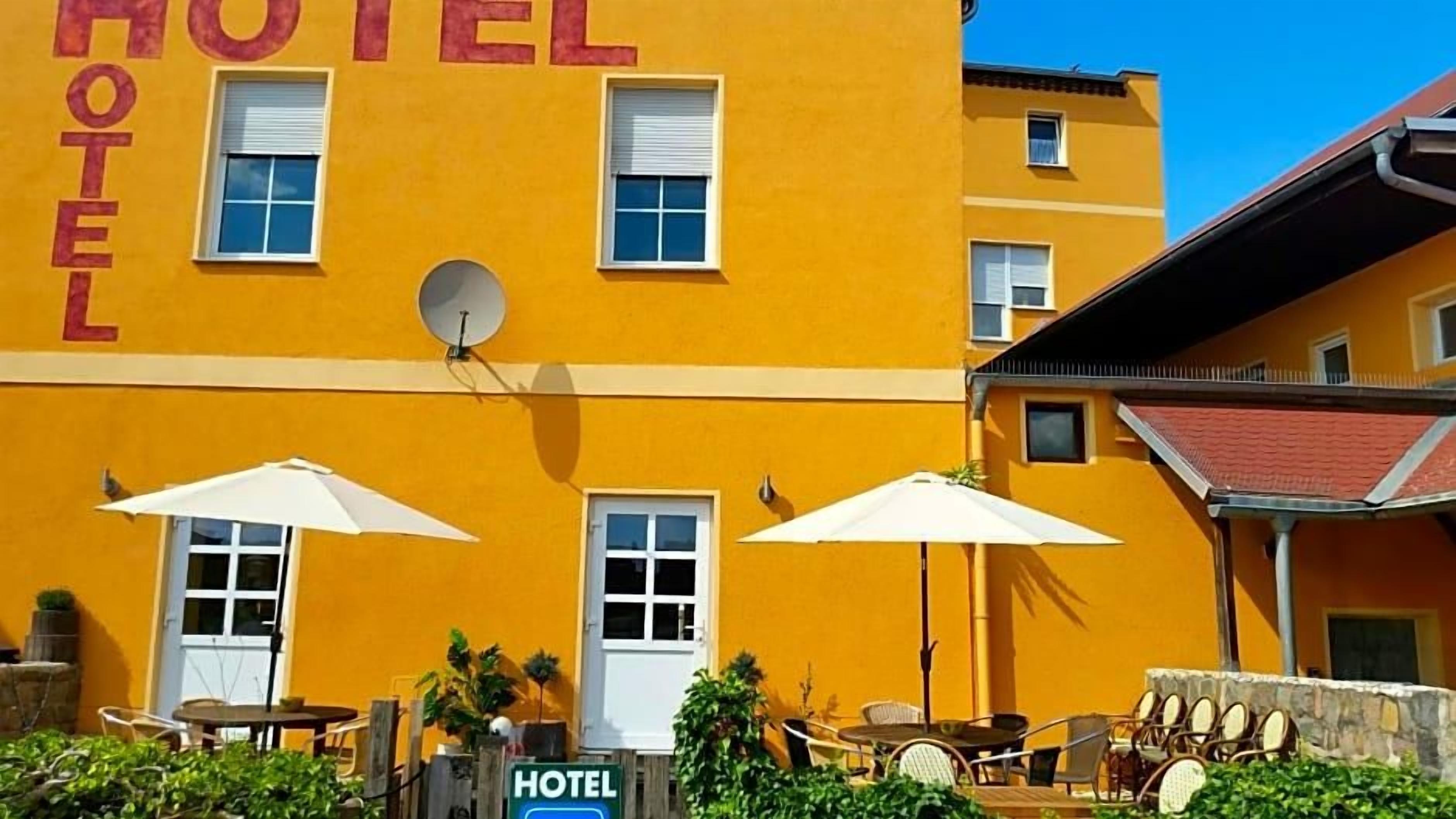 A yellow building with 'HOTEL' written on the facade, outdoor seating with umbrellas, and a sign indicating the hotel's location.
