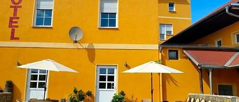 A yellow building with 'HOTEL' written on the facade, outdoor seating with umbrellas, and a sign indicating the hotel's location.
