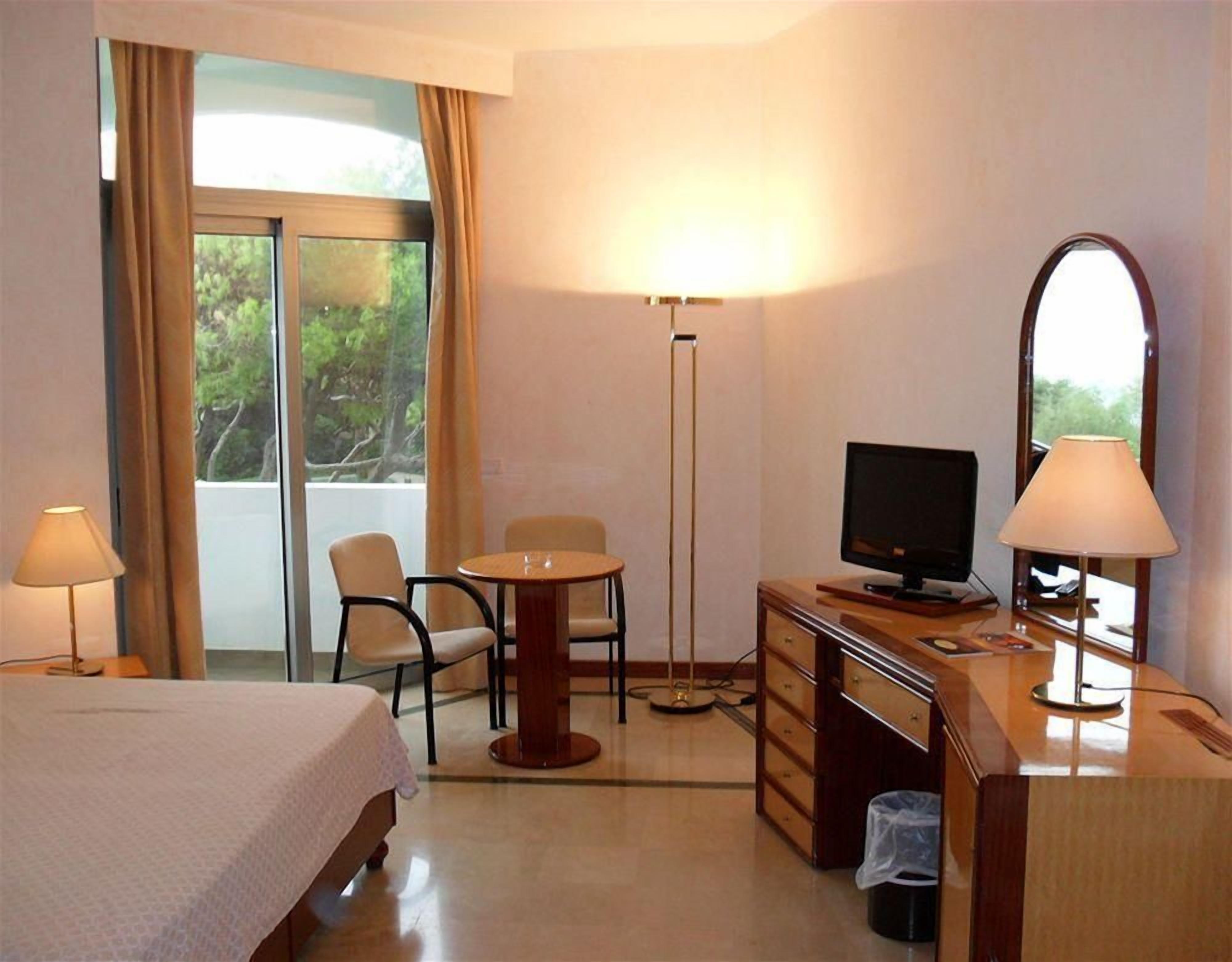 A hotel room with a bed, a desk with a TV, a chair, a small table, and a floor lamp.