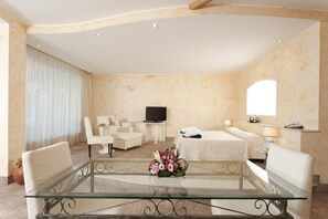 Une chambre d’hôtel avec une table basse en verre, deux fauteuils blancs, un lit avec une couette blanche, une télévision et un miroir au mur.