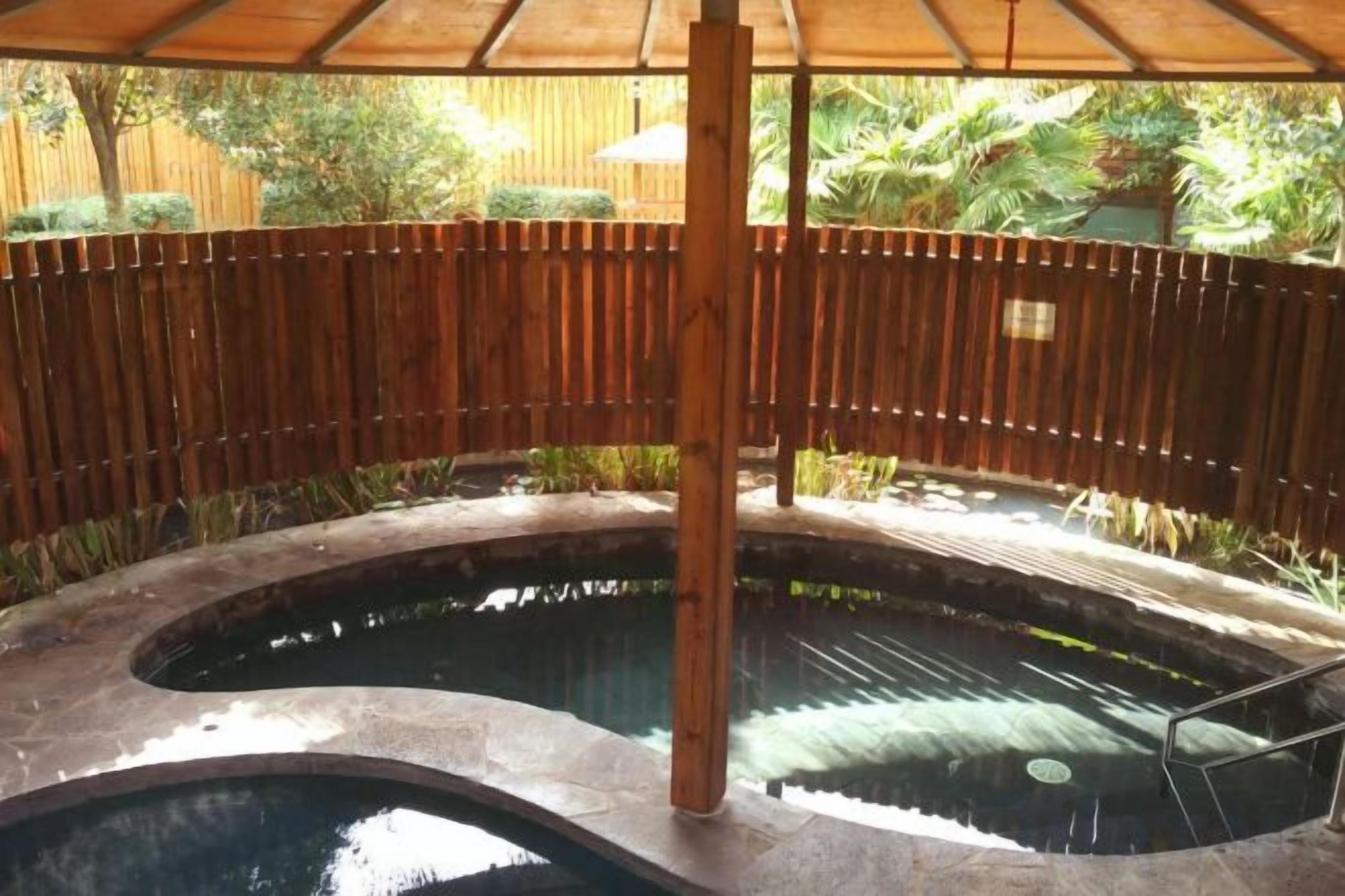 A circular pool with a wooden fence surrounding it, a wooden canopy overhead, and a view of greenery in the background.