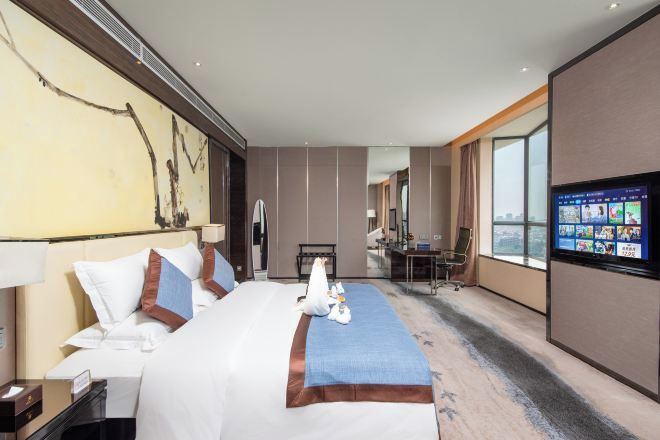 Ein Hotelzimmer mit einem großen Bett, einem Schreibtisch, einem Sessel und einem Fernseher, der verschiedene Sender anzeigt.