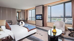 A hotel room with a large bed, a desk with a computer, a television, and a view of the city through large windows.