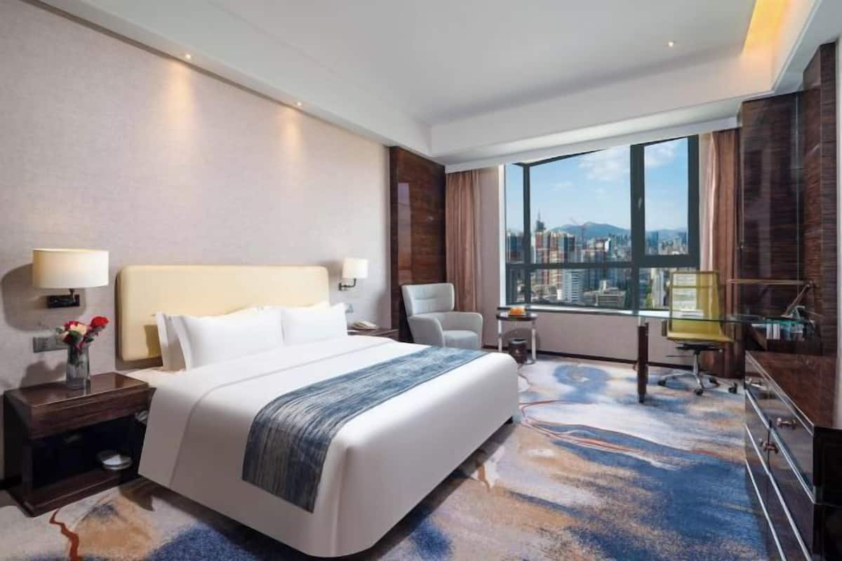 Une chambre d’hôtel avec un grand lit, un bureau, une chaise, une table de chevet et une vue sur le paysage urbain, visible à travers une grande fenêtre.