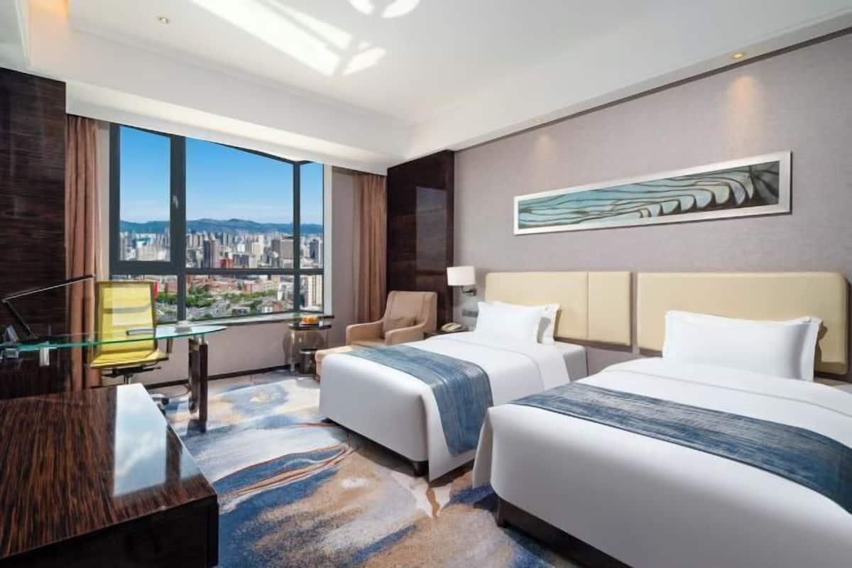 Une chambre d’hôtel avec deux lits, un bureau, une chaise, une grande fenêtre donnant sur la ville et un tableau au mur.