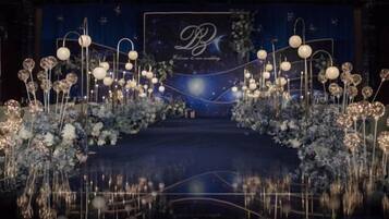 A stage with floral arrangements and lighting fixtures, a logo with 'B' and 'B' letters, and a dark background.