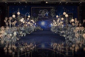 Un escenario con arreglos florales e iluminación, un logotipo con las letras 'B' y 'B', y un fondo oscuro.