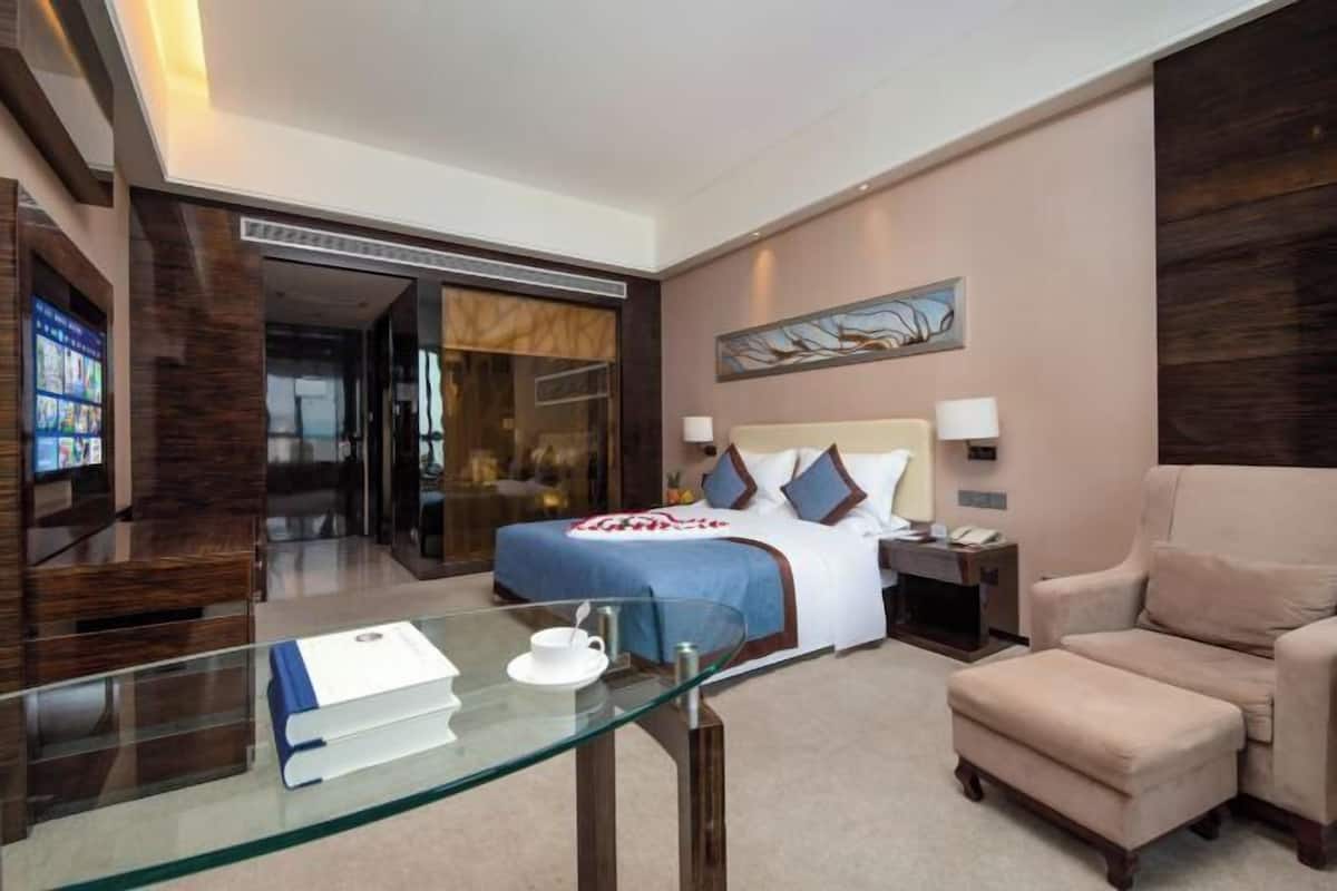 Une chambre d’hôtel avec un lit, une table en verre sur laquelle se trouvent un livre et une tasse, une chaise, une table de chevet et un tableau accroché au mur.
