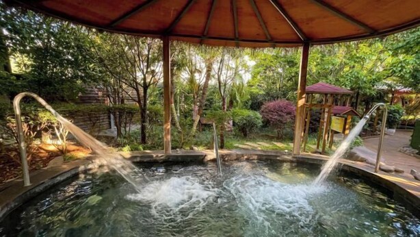 A hot spring area with two water jets, surrounded by lush greenery and a wooden gazebo.