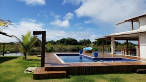 A swimming pool with a wooden deck, a covered patio area, and a view of a lake.