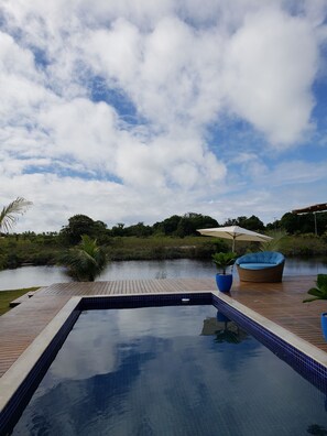 A swimming pool with a wooden deck, surrounded by lush greenery and a calm body of water.