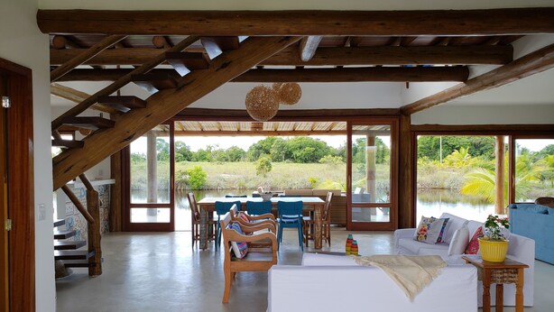 A spacious room with a high ceiling, wooden beams, a dining area with blue chairs, a white sofa, and a view of a natural landscape through large windows.