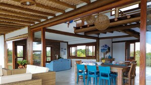 A spacious living area with a dining table and blue chairs, a wicker sofa, and a large window offering a view of greenery.