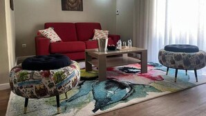 Een woonkamer met een rode bank, twee voetenbankjes, een salontafel en een kleurrijk vloerkleed.