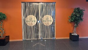 Een receptiegedeelte met een houten deur waarop de naam 'OLIVE' staat, en een bord erboven. Aan weerszijden van de deur staan twee potplanten.