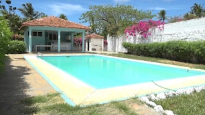 Piscina com deck revestido em azulejo, área de estar coberta e parede branca com flores cor-de-rosa.