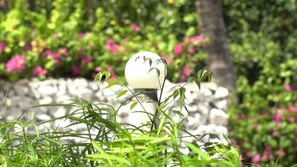 A garden with a spherical white object, possibly a sculpture, surrounded by green plants and pink flowers.