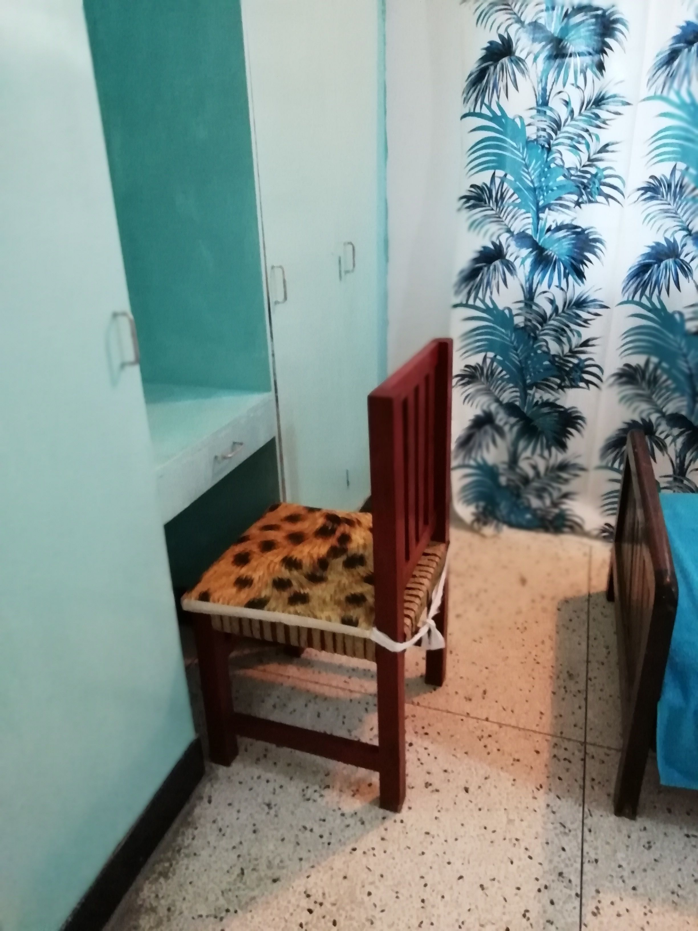 A corner room with a wooden chair with a leopard print cushion, a blue curtain with palm tree patterns, and a tiled floor.