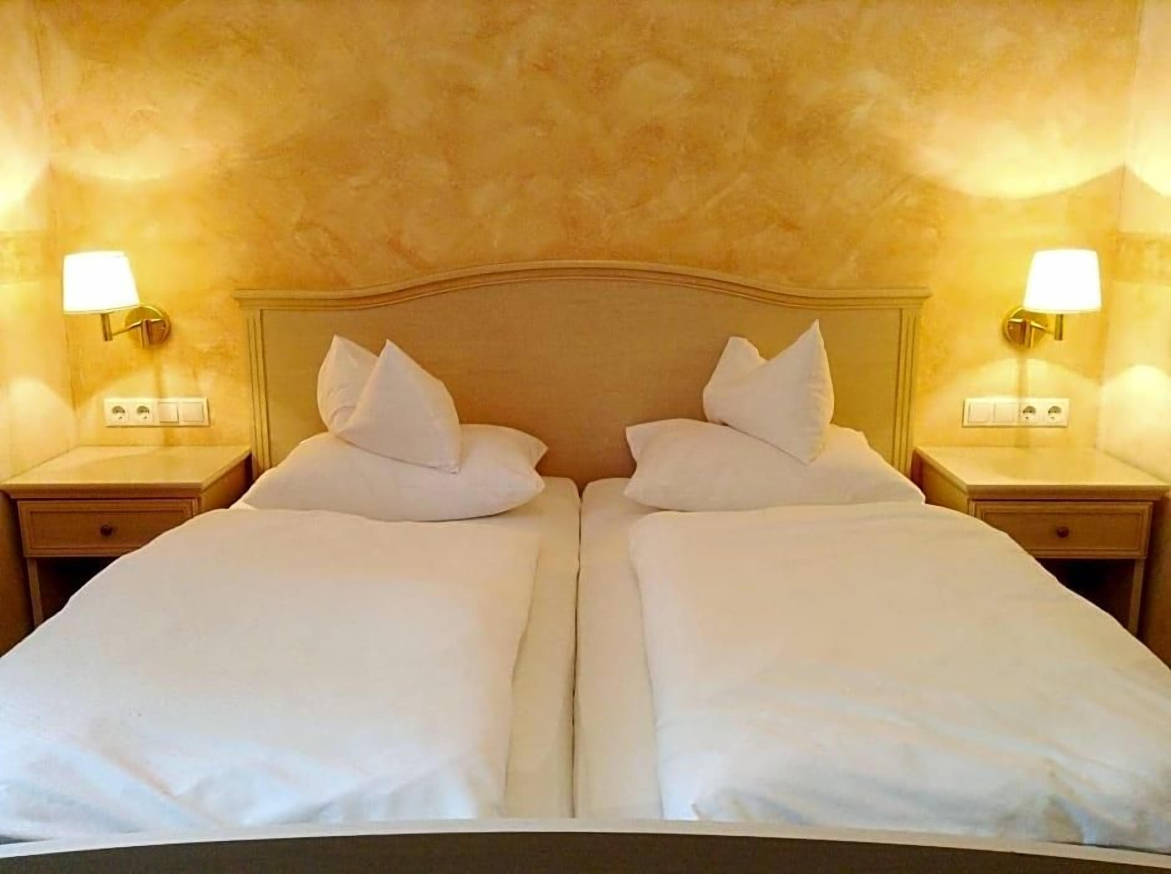 A double bed with white linens and pillows, flanked by two bedside tables with lamps, in a room with textured walls.