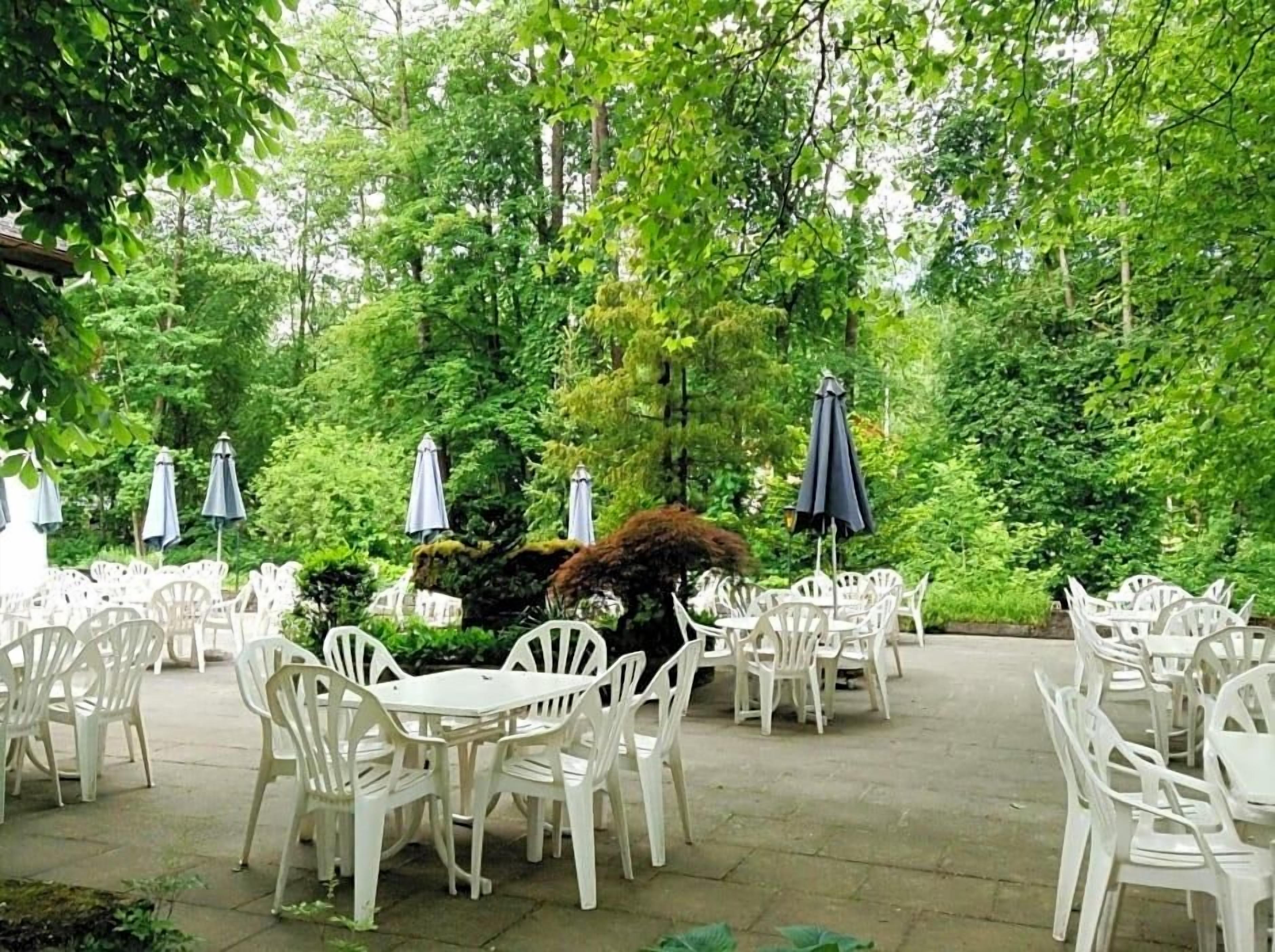 An outdoor patio area with white plastic chairs and tables, surrounded by lush greenery and trees.