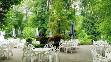 An outdoor patio area with white plastic chairs and tables, surrounded by lush greenery and trees.