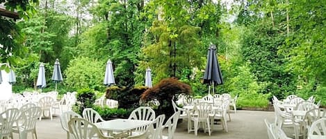 Un área de patio al aire libre con sillas y mesas de plástico blanco, rodeada de exuberante follaje y árboles.