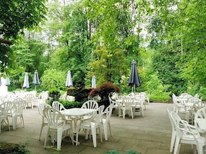 An outdoor patio area with white plastic chairs and tables, surrounded by lush greenery and trees.