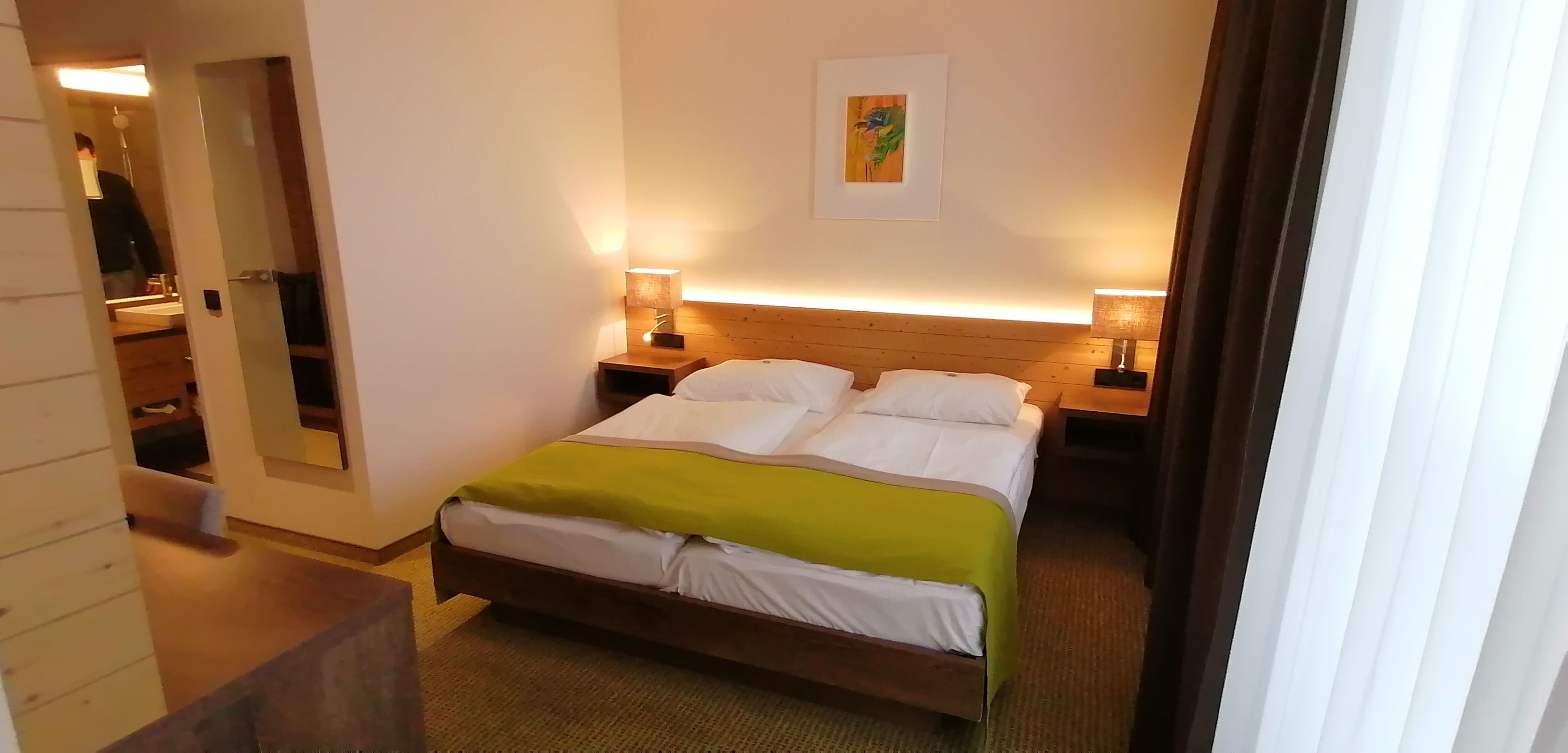 Una habitación de hotel con una cama, dos lámparas de noche, un cabecero de madera, una mesita de noche pequeña y un espejo.