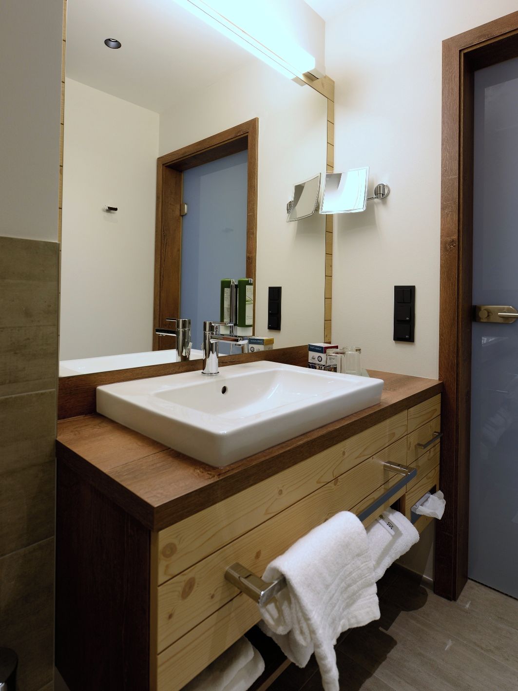 A modern bathroom with a large mirror, two sinks, wooden cabinetry, and a towel rack.