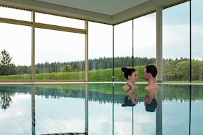Una pareja en una piscina interior con amplios ventanales y vistas a un paisaje boscoso.