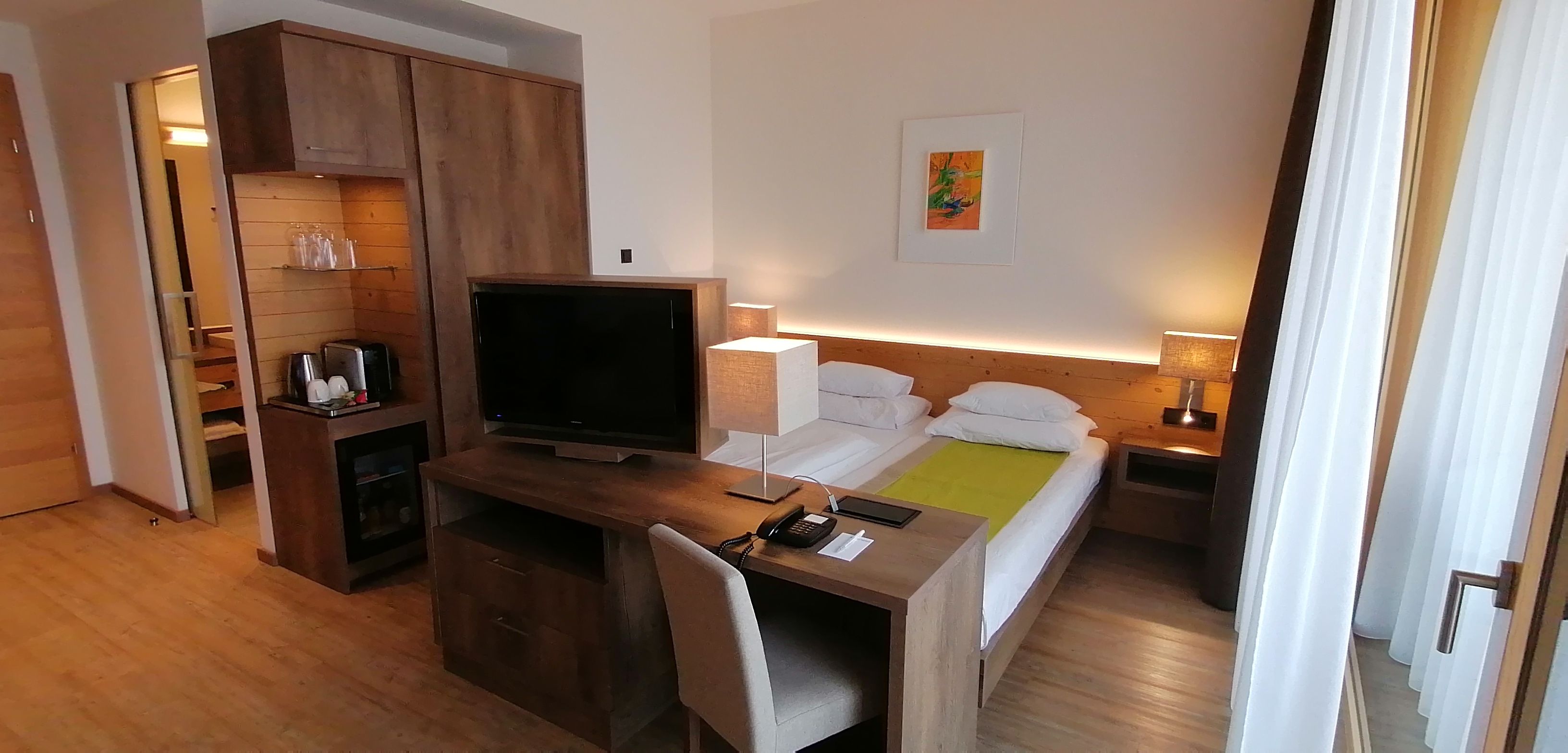 Ein Hotelzimmer mit einem Holztisch, einem Fernseher, einem Bett mit weißer Bettwäsche, einem Nachttisch mit einer Lampe und einem Bild an der Wand.