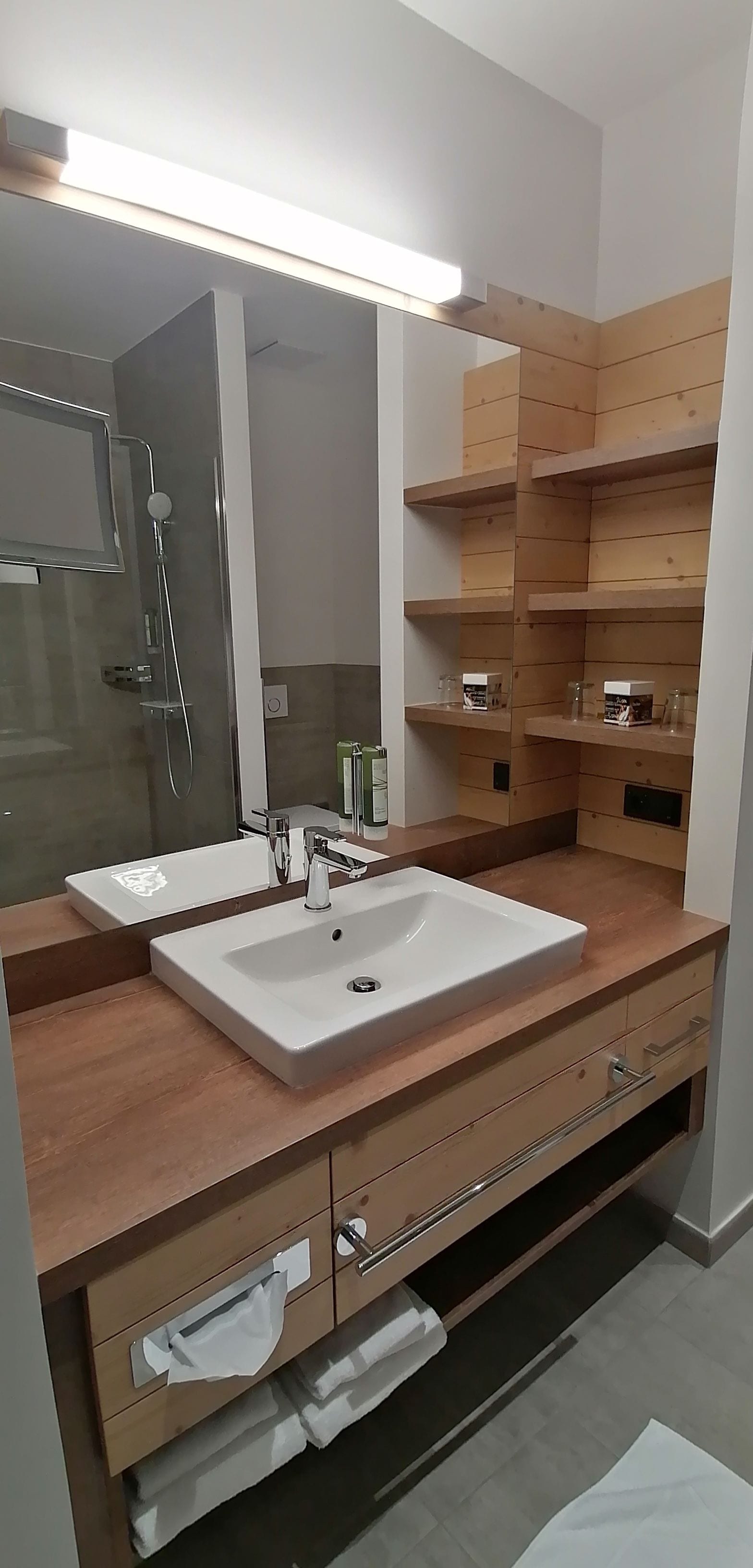 A modern bathroom with a large mirror, two sinks, wooden shelves, and a shower area.