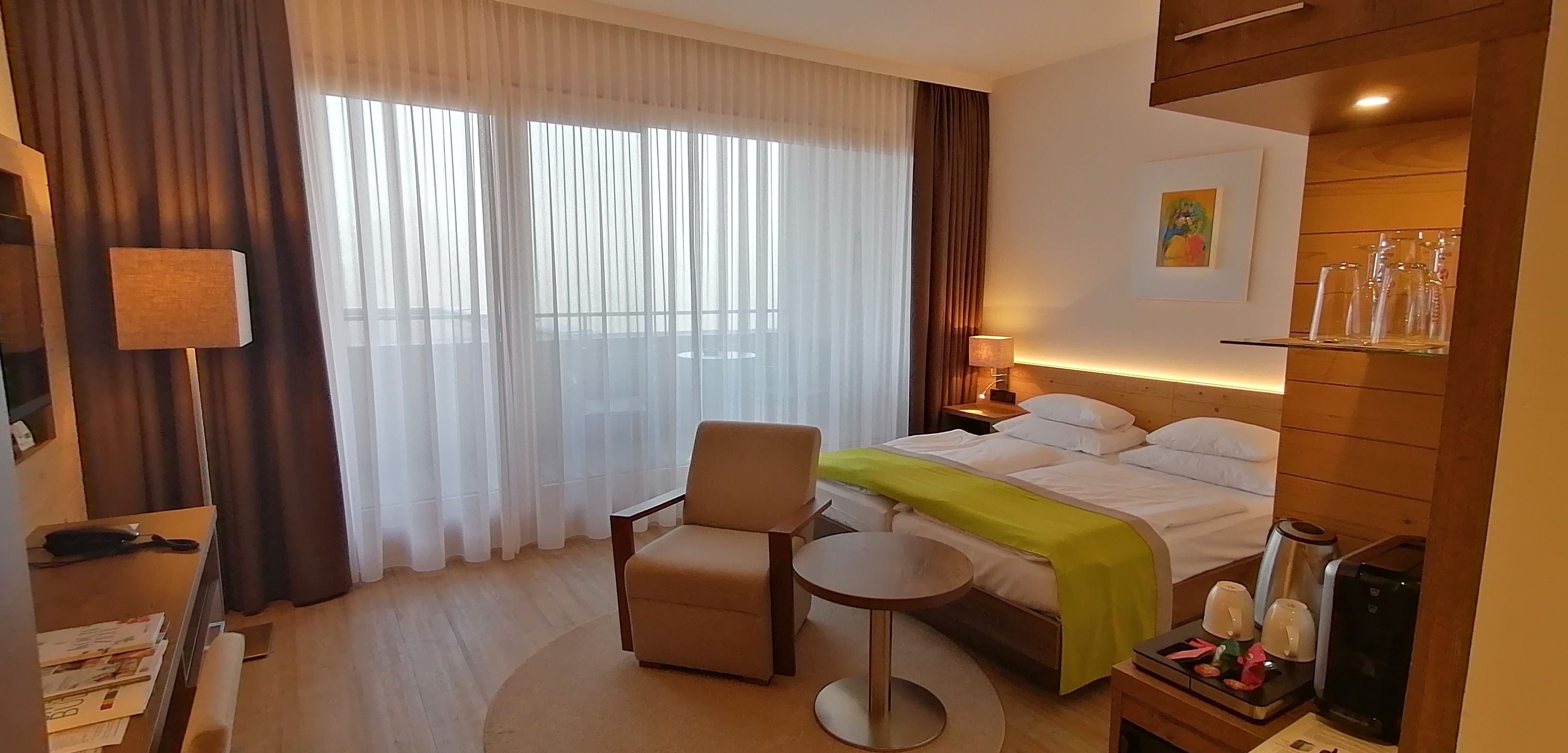 A hotel room with a bed, a chair, a small table, a lamp, and a nightstand with a lamp.