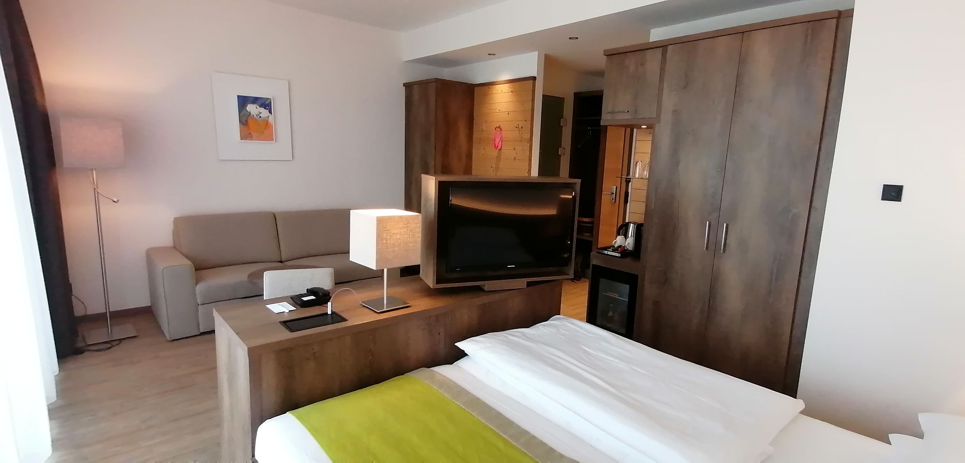 A hotel room with a bed, a desk with a TV, a sofa, and a wardrobe.