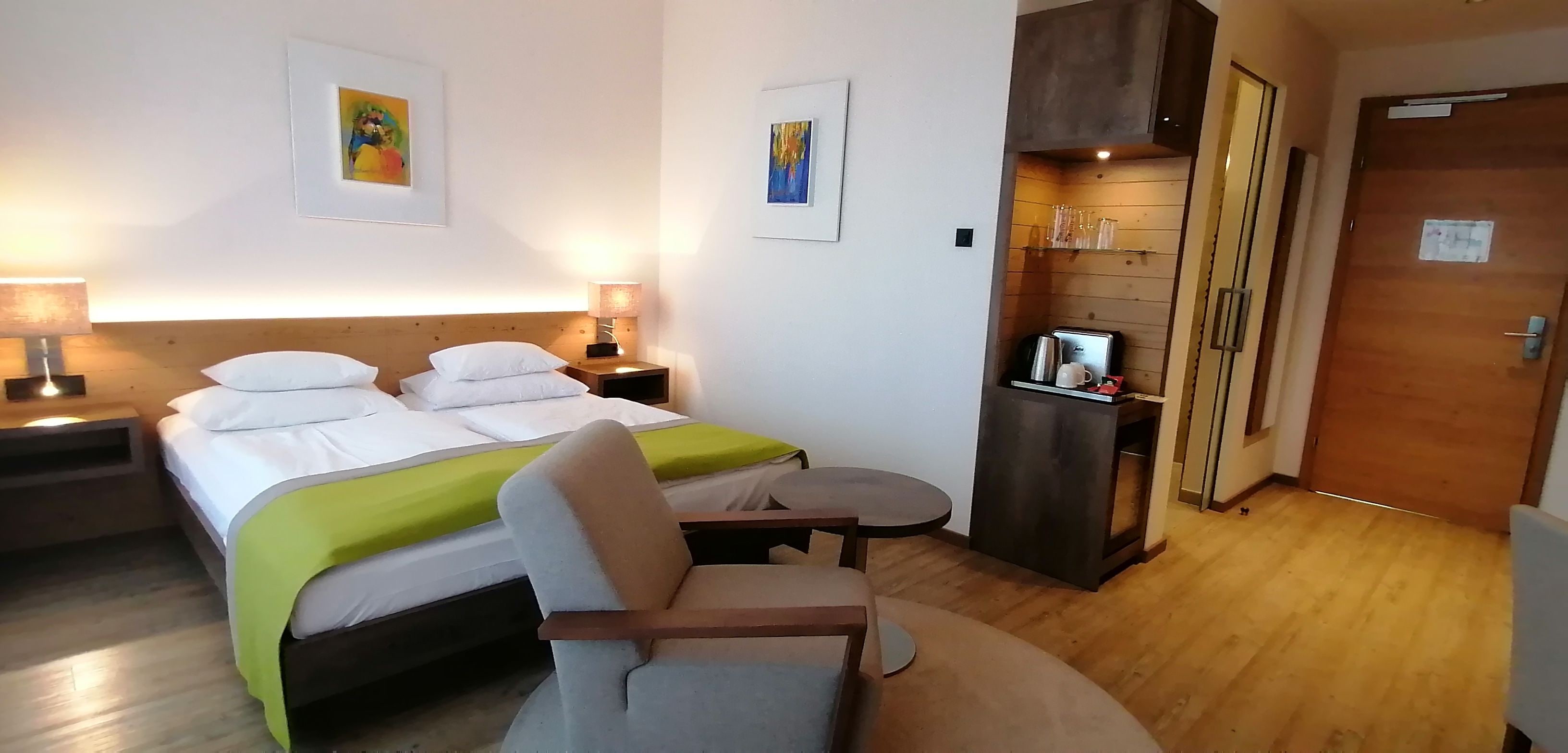 Habitació d'hotel amb llit, làmpades de nit, una cadira, una taula petita, un minibar i una porta.