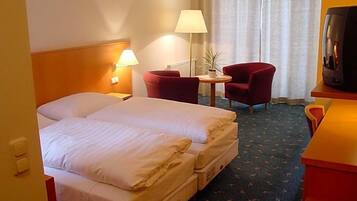 Ein Hotelzimmer mit einem Doppelbett, zwei roten Sesseln, einem kleinen Tisch mit Lampe sowie einem Fernseher auf einem Holzboden.