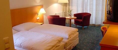 Ein Hotelzimmer mit einem Doppelbett, zwei roten Sesseln, einem kleinen Tisch mit Lampe sowie einem Fernseher auf einem Holzboden.