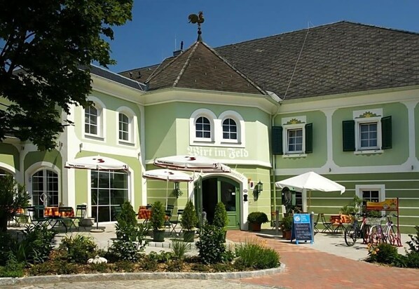 A building with a green facade, a central entrance, outdoor seating with umbrellas, and bicycles parked outside.