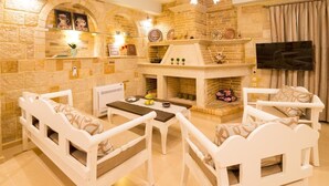 A cozy living room with a stone fireplace, white furniture, and a flat-screen TV. - Nikolas Villas Kamilari (Faistos)