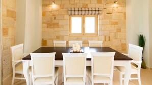 A dining area with a stone wall, a window with blinds, and a table with chairs. - Nikolas Villas Kamilari (Faistos)