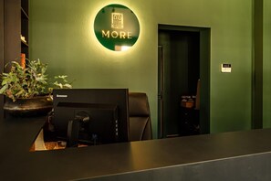 A reception desk with a Lenovo computer, a green wall with a 'MORE' sign, and a potted plant.