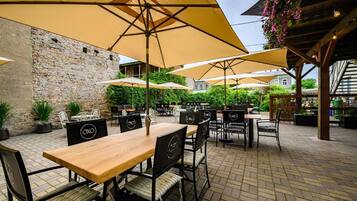 An outdoor patio with tables and chairs, covered by large umbrellas, and a stone wall in the background.