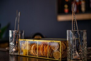 Un bar con una caja de vidrio que contiene rodajas de cítricos deshidratados, dos vasos con pajitas y una botella al fondo.