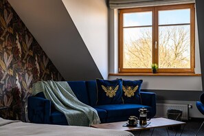 Una habitación con techo inclinado, un sofá azul con cojines decorativos, una mesita con cafetera y una ventana con vistas a árboles.